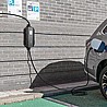 רכב חשמלי אפור מטעין ממטען לרכב חשמלי type 2 שחורה המותקנת על קיר לבנים אפור. הכבל השחור של המטען מחובר לשקע טעינה בצידו האחורי של הרכב. על הקיר משמאל לעמדת הטעינה תלוי שלט לבן עם האות